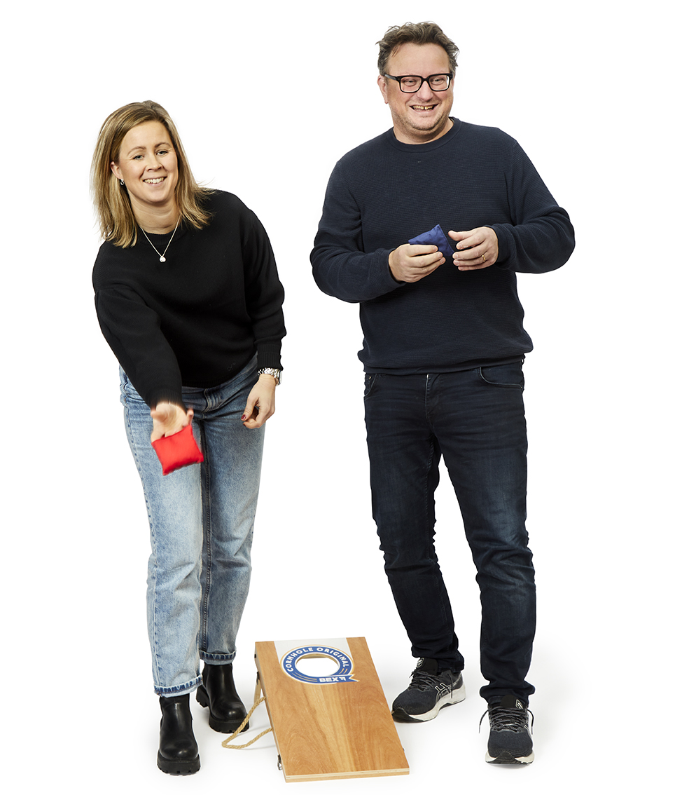Zwei Personen spielen Cornhole mit dem Produkt "Cornhole Original 60,5x30,7cm". Eine Frau wirft gerade ein rotes Säckchen auf das Holzbrett mit Loch, während ein Mann daneben steht und ein blaues Säckchen hält.
