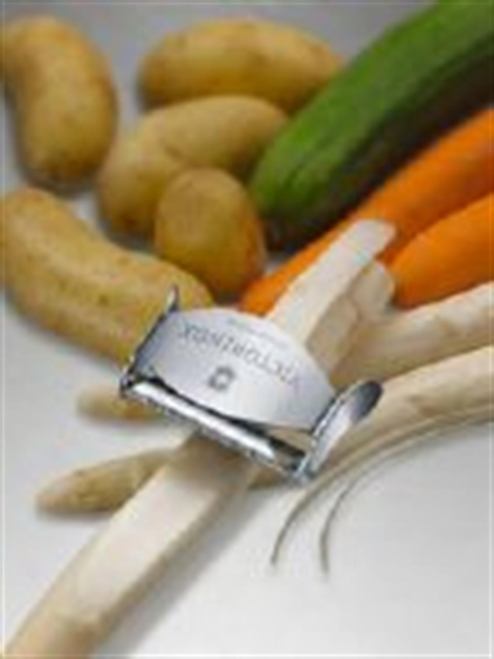 Auf dem Bild ist ein Sparschäler zu sehen, der gerade ein Gemüse schält. Im Hintergrund liegen verschiedene Gemüsesorten wie Kartoffeln, Karotten und eine Gurke.