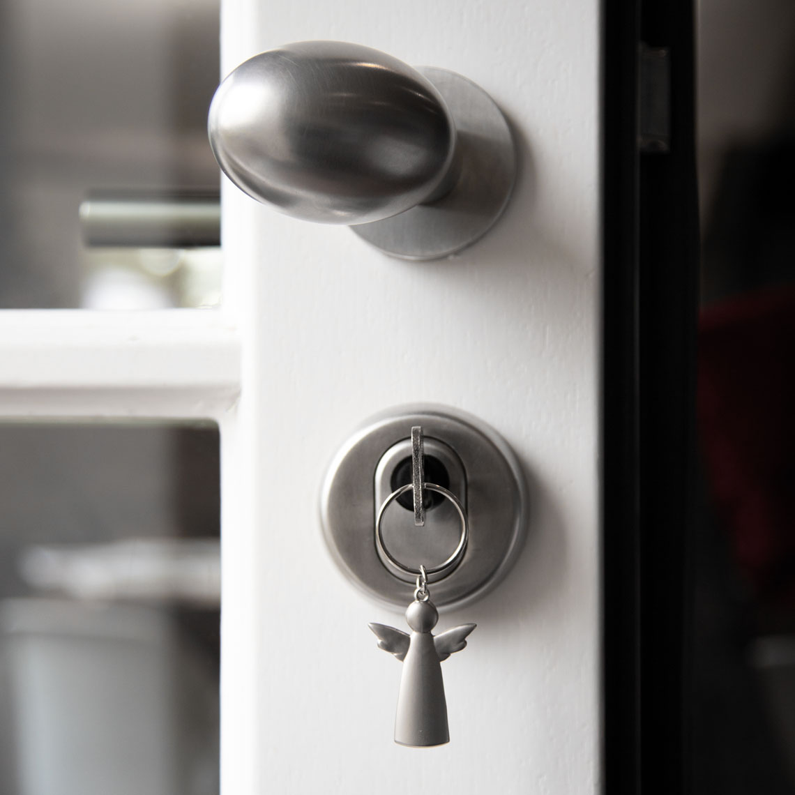 Auf dem Bild ist ein Türschloss mit einem silbernen Türgriff zu sehen. Im Schloss steckt ein Schlüsselbund mit einem Engel-Anhänger.