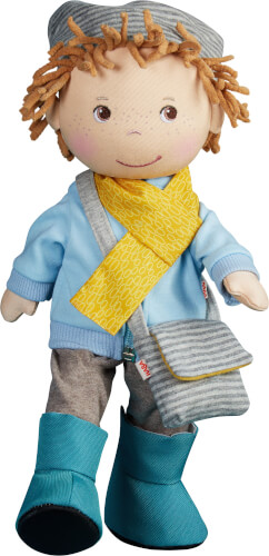Auf dem Bild ist eine Stoffpuppe mit braunen Wollhaaren und Sommersprossen zu sehen. Sie trägt eine blaue Jacke, eine gelbe Schal, eine graue Mütze, eine Umhängetasche, graue Hosen und blaue Stiefel.