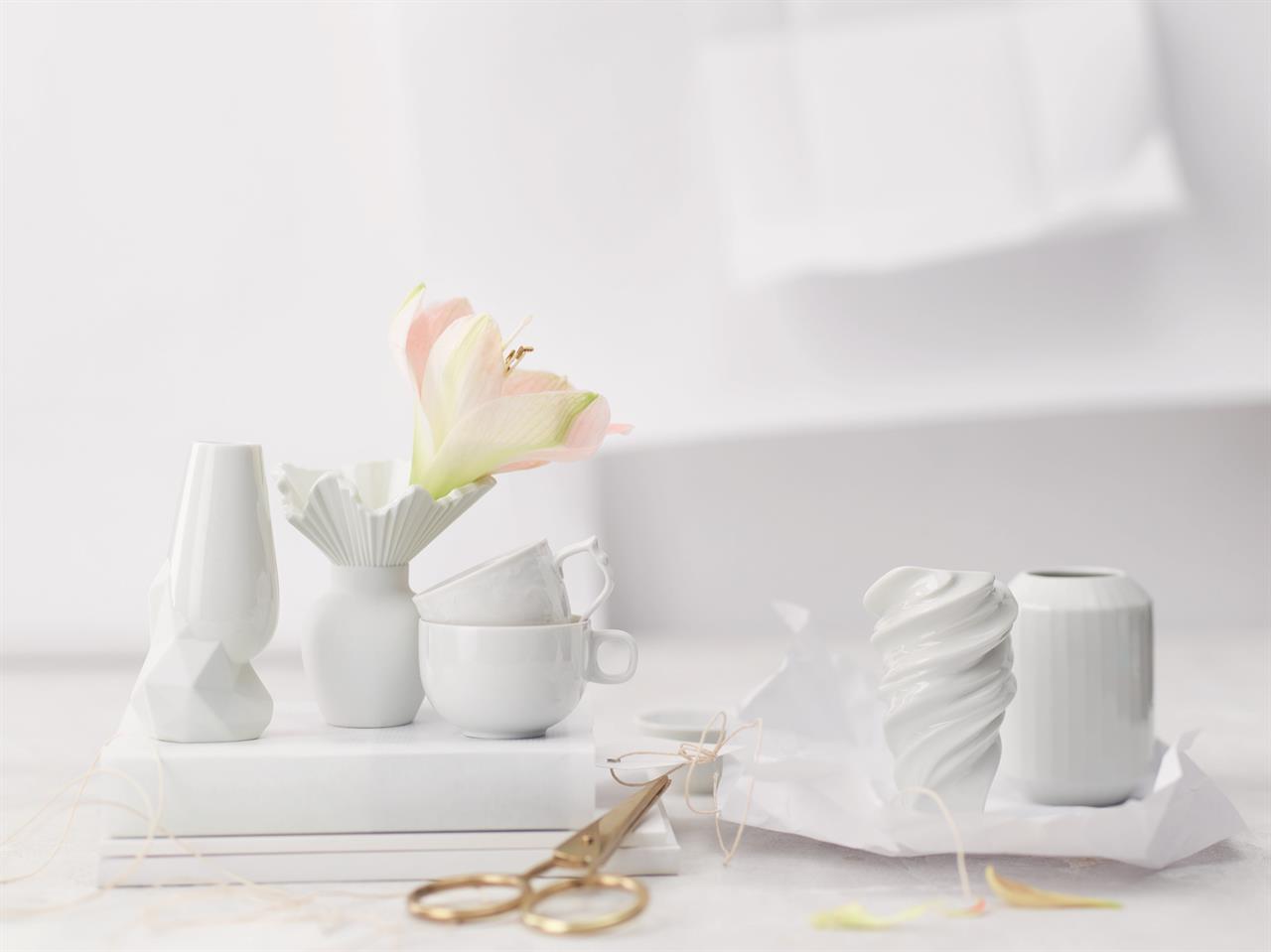 Auf dem Bild sind mehrere weiße Vasen und Tassen auf einem hellen Hintergrund zu sehen. Im Vordergrund steht die Vase "Squall / Weiß" mit einem gedrehten Design.