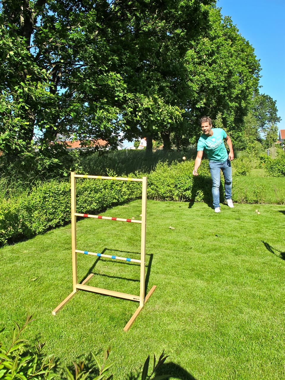 Ein Mann spielt im Garten mit dem Spiel "Spin Ladder Original - Leitergolf". Er wirft eine Bola auf das Holzgestell mit bunten Stangen.