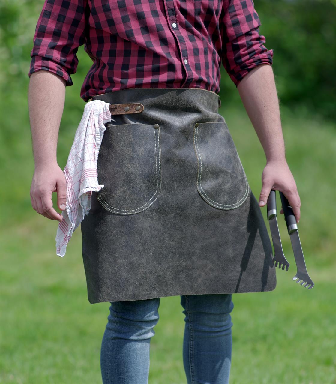 Eine Person trägt eine anthrazitfarbene Hüftschürze aus Antikleder in der Größe 70x55 cm. In der Schürze stecken ein Geschirrtuch und Grillbesteck.