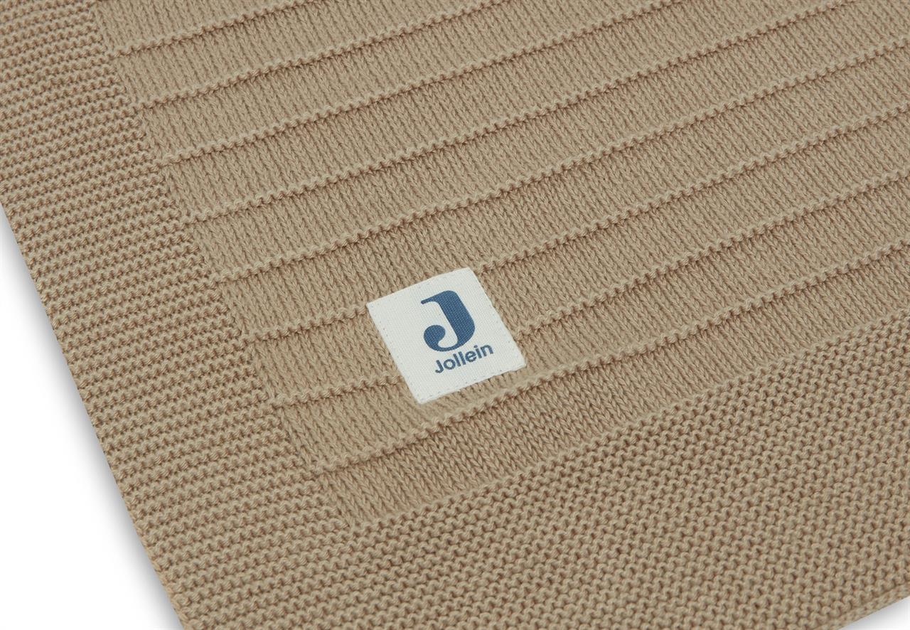 Auf dem Bild ist eine beige gestrickte Decke mit einem Etikett der Marke Jollein zu sehen. Die Decke hat ein geripptes Strickmuster und eine abgesetzte Umrandung.