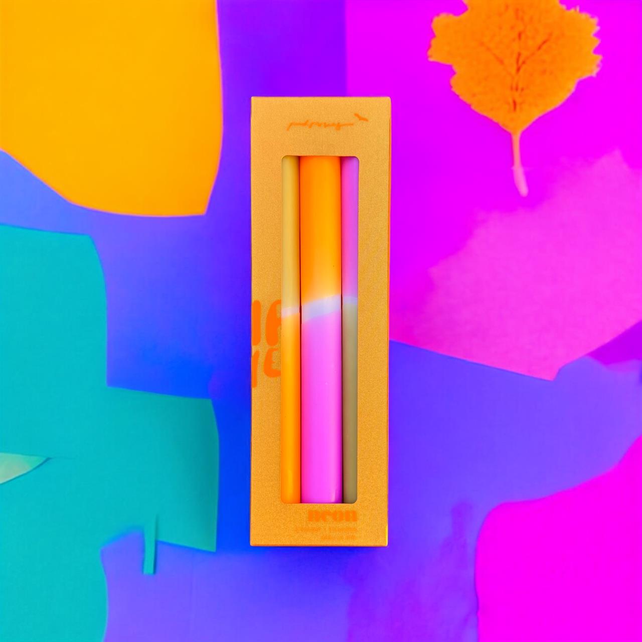 Das Bild zeigt das Produkt "Kerzen Dip Dye Neon 'Honey Glaze'" in einer orangenen Verpackung. Der Hintergrund ist bunt und leuchtend mit Farben wie Pink, Lila, Gelb und Türkis gestaltet.