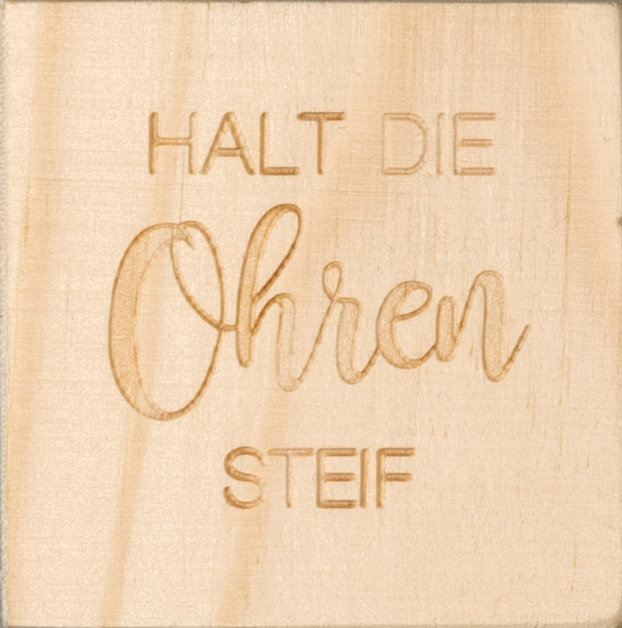 Auf dem Bild ist eine Holzplatte mit einer Gravur zu sehen. Der Text lautet: "HALT DIE Ohren STEIF".