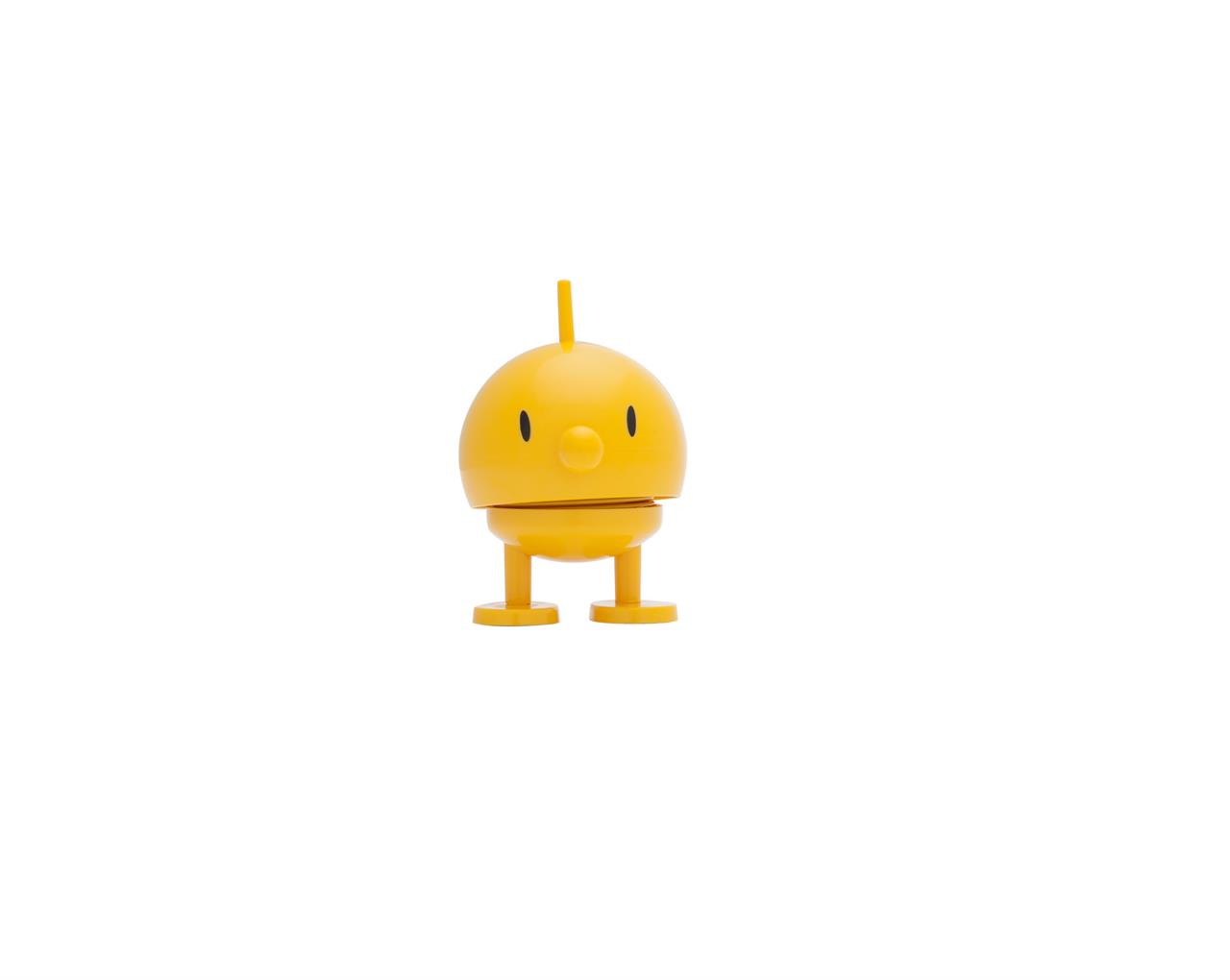 Das Bild zeigt eine gelbe, runde Figur mit zwei Beinen und einem kleinen Stab auf dem Kopf. Die Figur hat ein freundliches Gesicht und steht auf weißem Hintergrund.