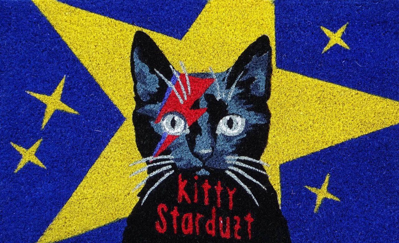 Das Foto zeigt das Produkt 'Fussmatte, Motiv: Kitty Starduzt, blau/gelb'. Die Fußmatte ist mit einer stilisierten Katze in Blau und Gelb verziert.