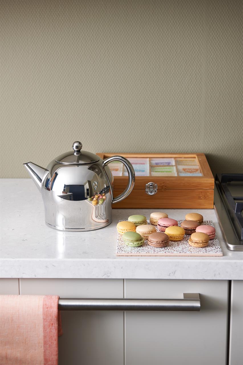 Auf dem Bild ist eine glänzende Teekanne „Teekanne Minuett Santhee 1.4L“ auf einer Küchenarbeitsplatte zu sehen. Daneben stehen ein Tablett mit bunten Macarons und eine Holzkiste mit Teebeuteln.