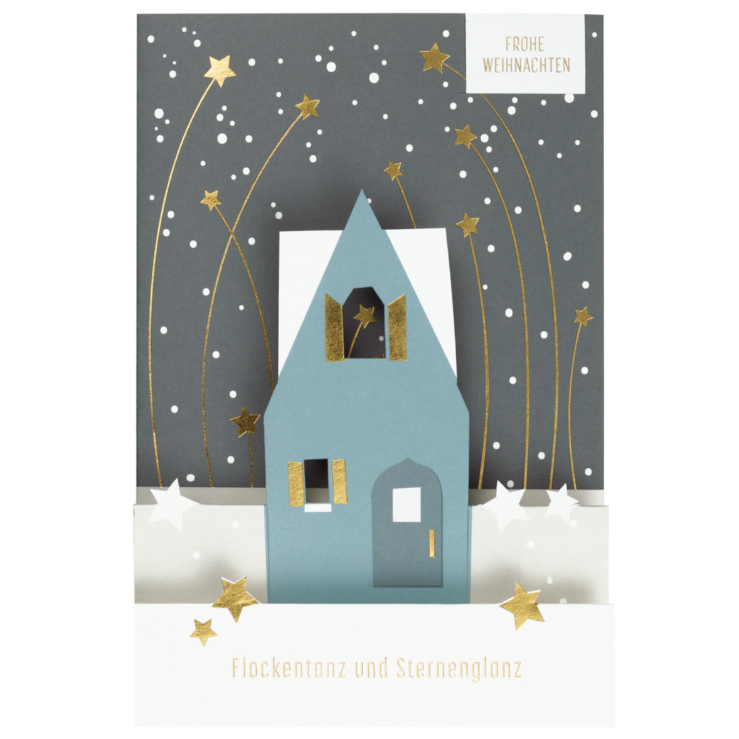 Auf dem Bild ist eine Weihnachtskarte mit einem 3D-Papierhaus vor einem winterlichen Nachthimmel zu sehen. Goldene Sterne und Schneeflocken schmücken die Karte, auf der "Flockentanz und Sternenglanz" steht.
