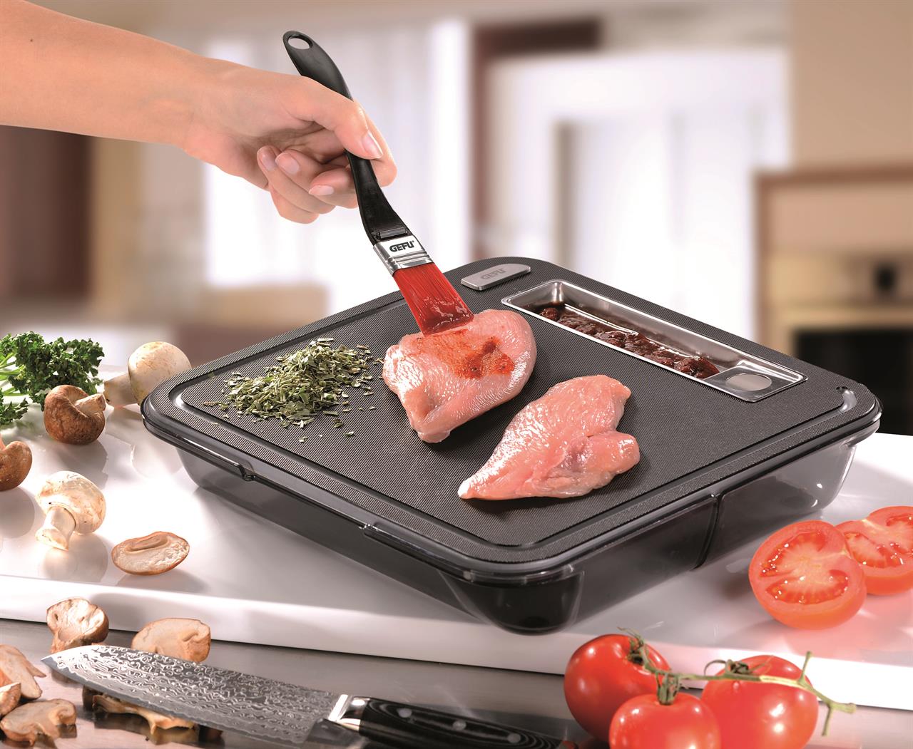 Eine Hand bestreicht mit dem Küchenpinsel Becky ein Stück rohes Hähnchenfleisch mit Marinade. Auf dem Schneidebrett liegen außerdem Kräuter, Champignons und Tomaten.