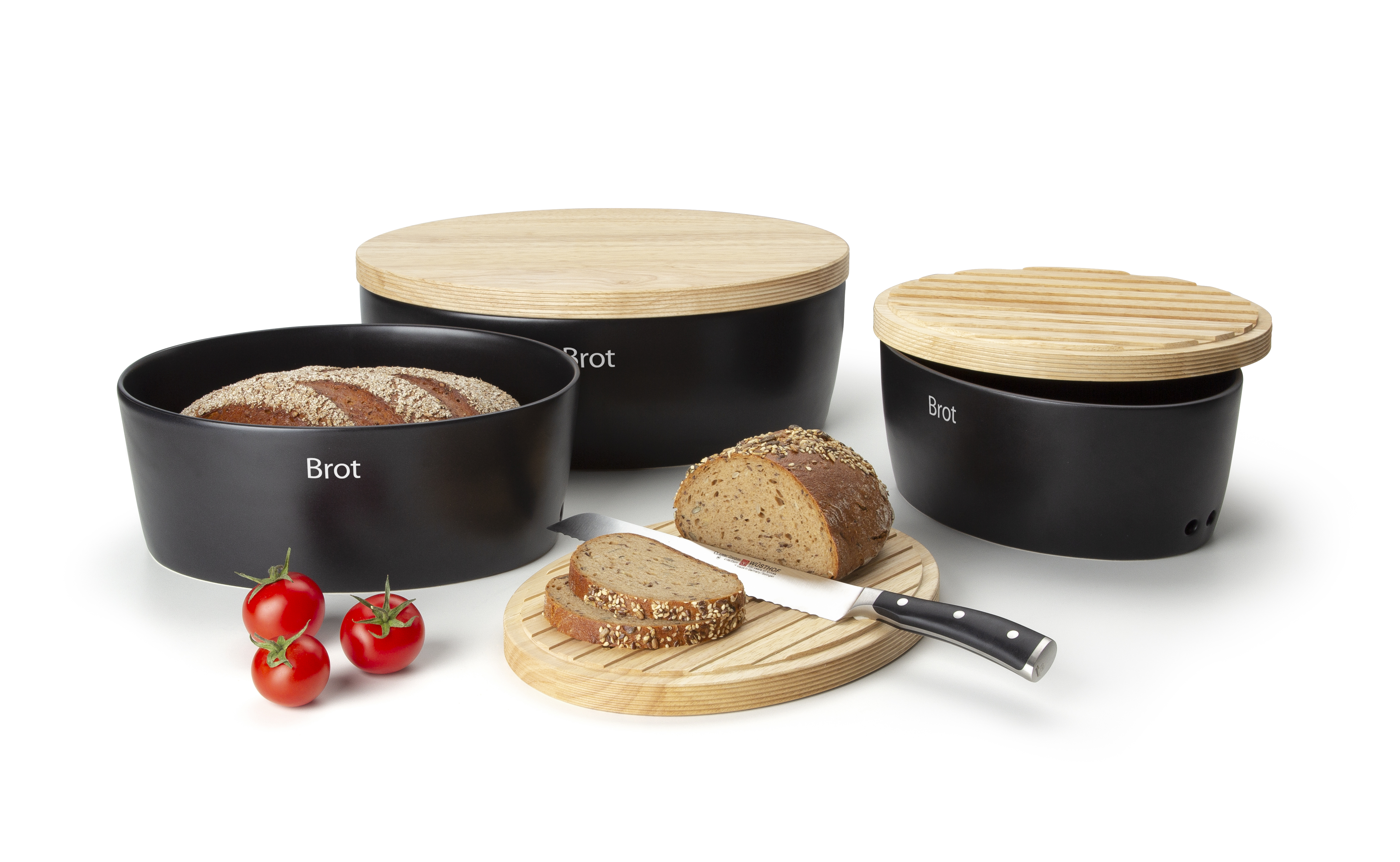 Auf dem Bild sind drei schwarze Brotdosen mit Holzdeckeln zu sehen, auf denen "Brot" steht. Davor liegen geschnittenes Brot auf einem Holzbrett, ein Messer und drei Tomaten.