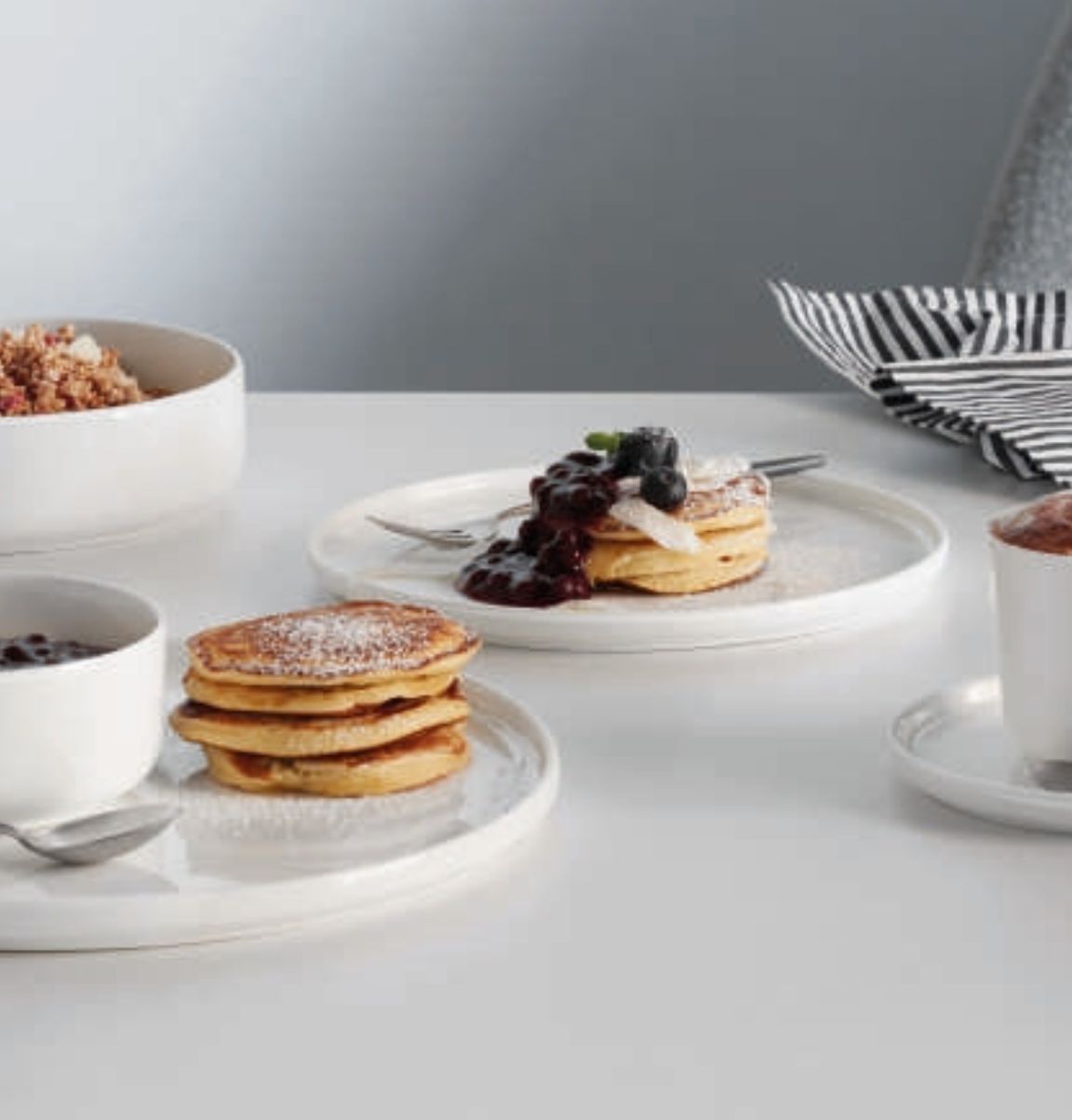 Auf dem Tisch stehen Teller mit Pancakes, die mit Puderzucker und Heidelbeersoße garniert sind. Daneben befinden sich eine Schale mit Müsli, eine Tasse und gestreifte Servietten.