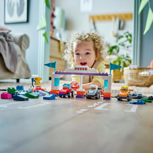 Ein kleines Kind spielt auf dem Boden mit bunten Lego-Autos und Bauklötzen. Im Hintergrund ist ein gemütlich eingerichtetes Wohnzimmer zu sehen.