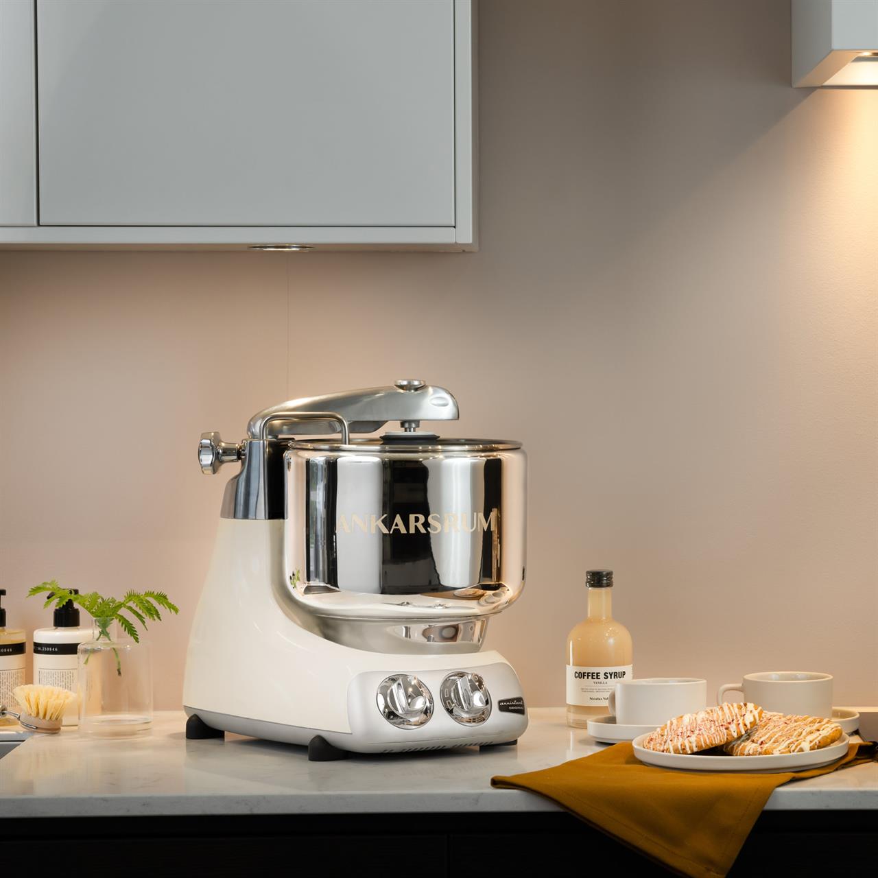 Die Küchenmaschine Light Creme steht auf einer Küchenarbeitsplatte. Daneben stehen Tassen, Gebäck und eine Flasche Kaffeesirup.