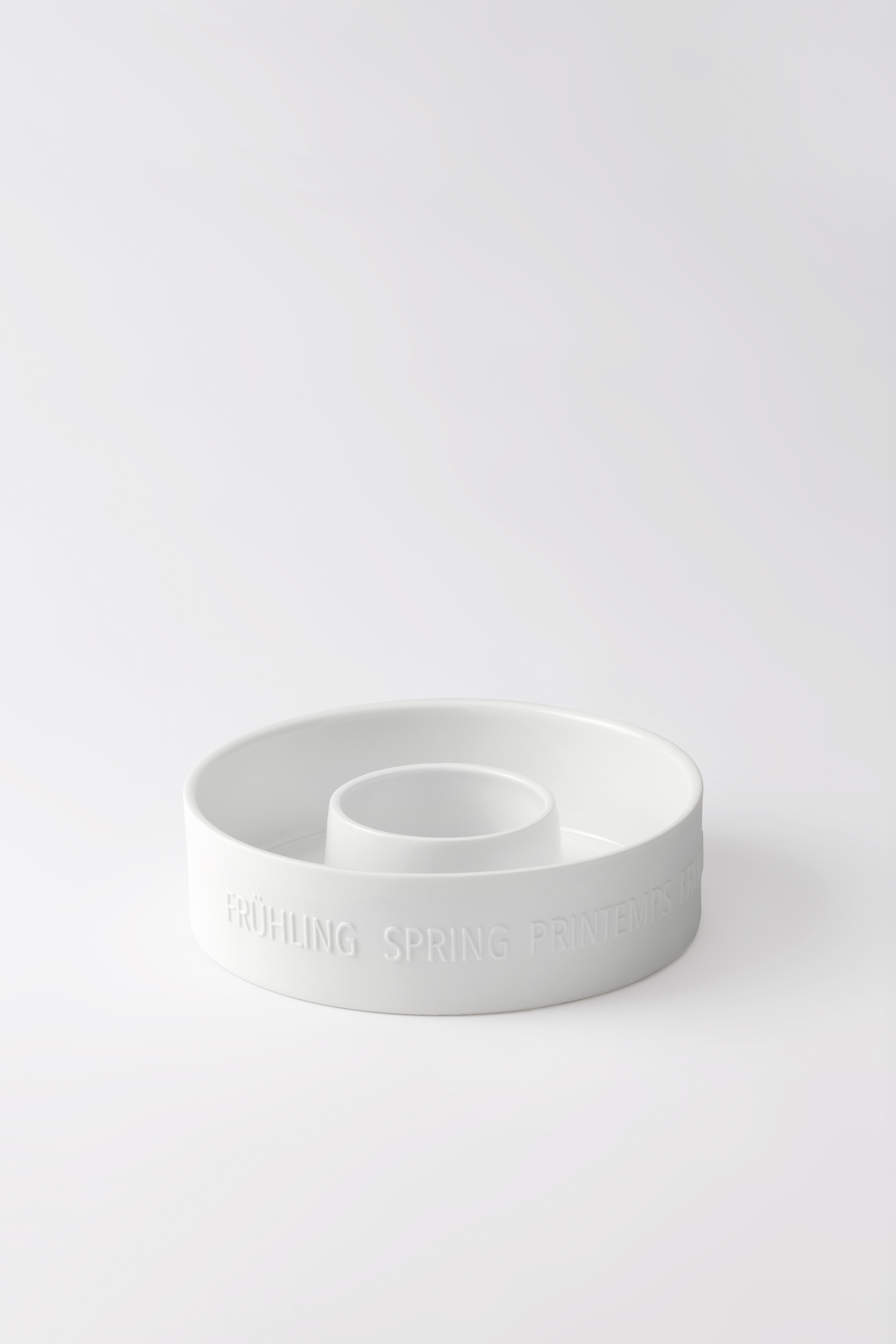 Das Bild zeigt eine runde, weiße Schale mit einem erhöhten Ring in der Mitte. Am Rand der Schale ist das Wort "FRÜHLING" zusammen mit seinen Übersetzungen geprägt.
