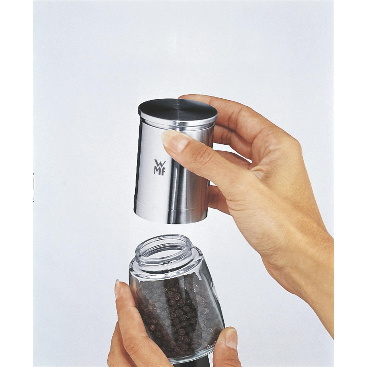 Auf dem Bild ist die Gewürzmühle De Luxe Unbefüllt zu sehen. Eine Hand hält das Edelstahl-Oberteil, während die andere ein Glas mit Pfefferkörnern darunter hält.