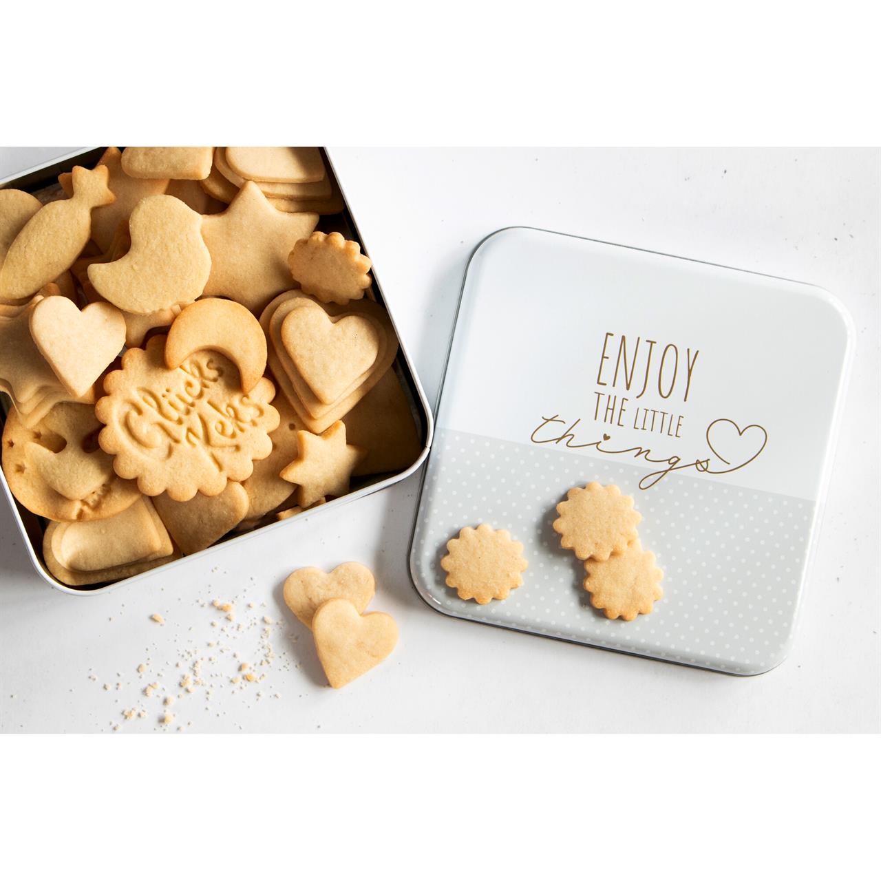 Die quadratische Gebäckdose mit der Aufschrift „Enjoy the little things“ ist mit verschiedenen Keksen gefüllt. Der Deckel liegt daneben, und einige Kekse sind auf dem Deckel und dem Tisch verstreut.