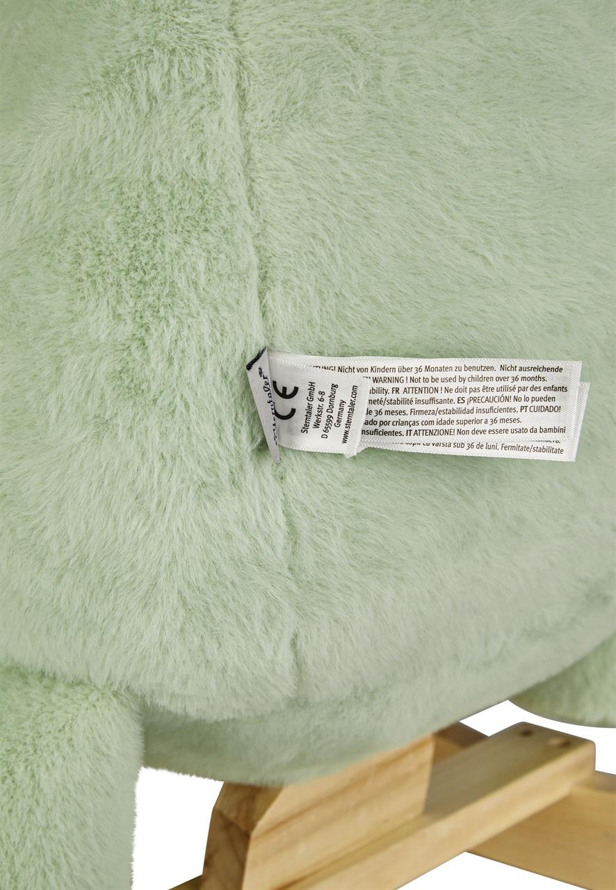 Das Bild zeigt einen Teil des Produkts "Schaukeltier Dino Rexi" in pudergrüner Farbe. Man sieht das flauschige Plüschtier und einen Holzsockel sowie ein Etikett mit Warnhinweisen.