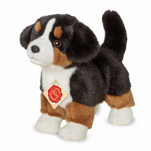 Das Bild zeigt ein Plüschtier in Form eines Hundes mit schwarzem, braunem und weißem Fell. Es trägt ein rotes Etikett um den Hals.
