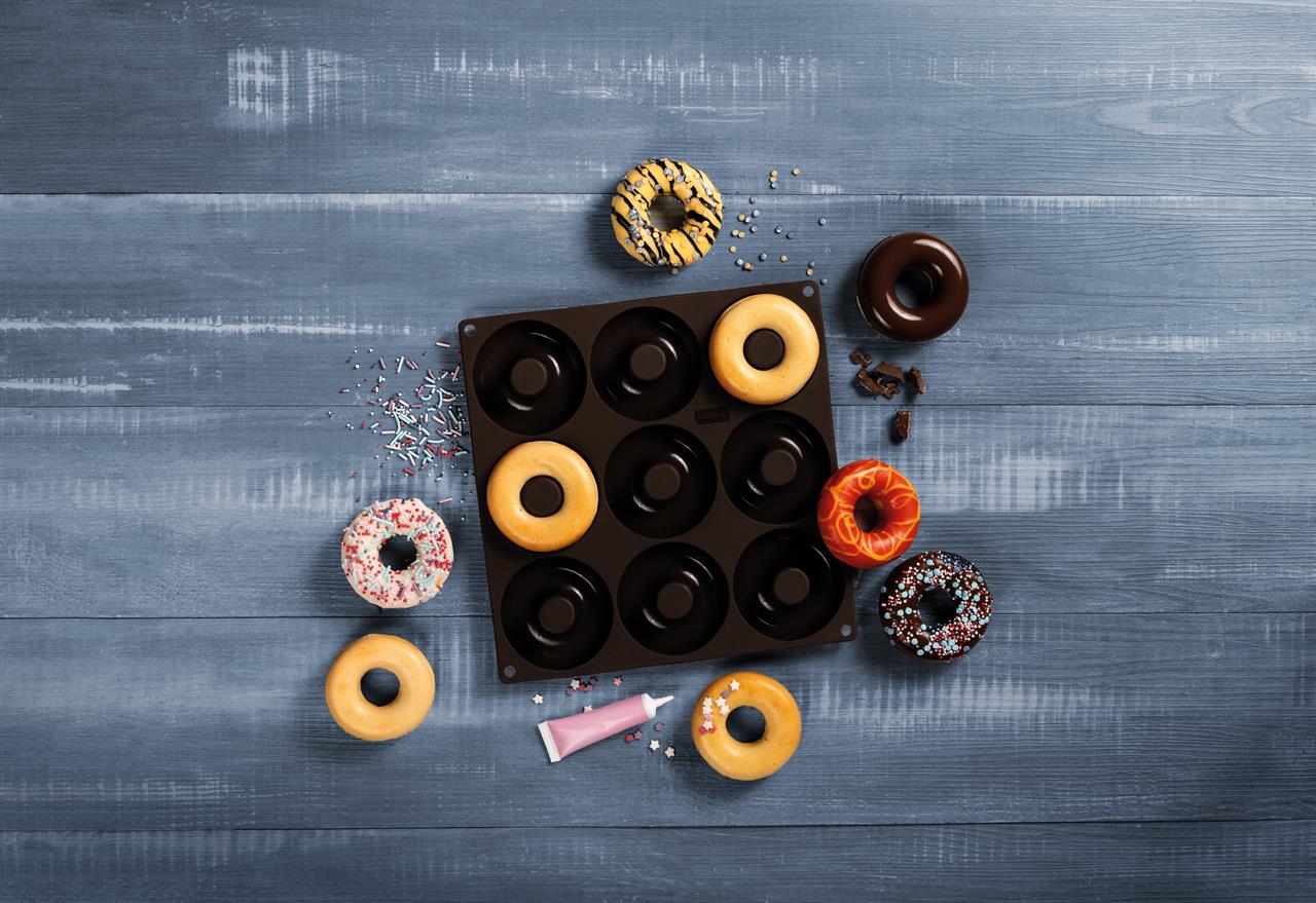 Das Bild zeigt eine braune Flexiform Donuts Backform mit neun runden Mulden auf einem blauen Holztisch. Um die Backform liegen mehrere glasierte und dekorierte Donuts sowie Streusel und eine Tube Zuckerguss.