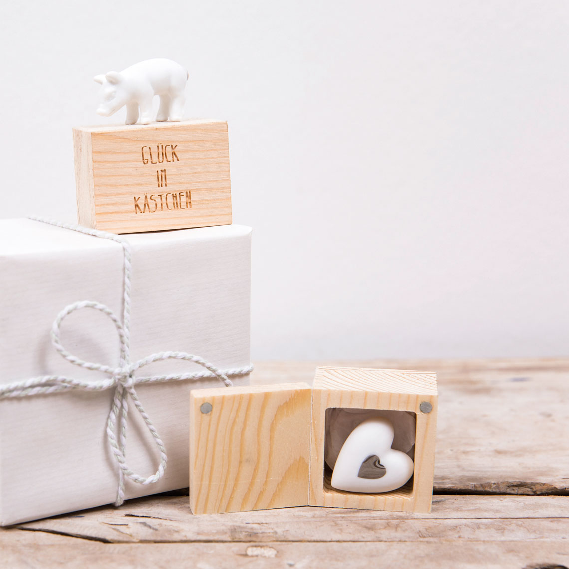 Auf dem Bild ist ein kleines Holzkästchen mit einer weißen Porzellanfigur eines Schweins zu sehen. Daneben steht eine weitere Holzdose mit einem Herz darin, und im Hintergrund liegt ein in weißes Papier eingepacktes Geschenk.