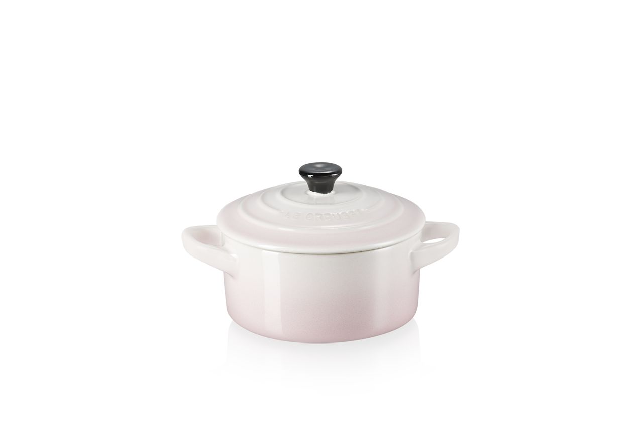 Das Bild zeigt eine kleine, runde Cocotte in sanftem Rosa mit zwei Henkeln und Deckel. Der Deckel hat einen schwarzen Griff und das Gefäß steht auf einer weißen Fläche.