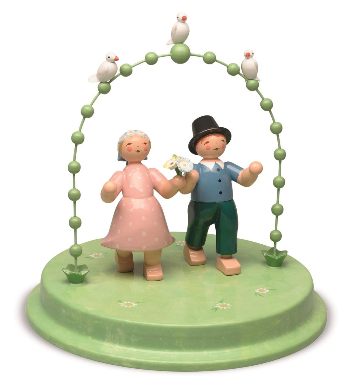 Das Bild zeigt ein Liebespaar aus Holzfiguren, das unter einem grünen Bogen steht. Auf dem Bogen sitzen drei weiße Täubchen.
