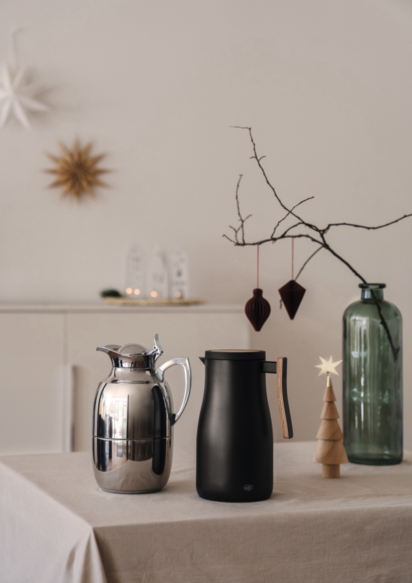 Auf dem Tisch stehen eine silberne Thermoskanne und eine schwarze Kanne nebeneinander. Im Hintergrund sind weihnachtliche Dekorationen wie Holzfiguren, ein großer Glasbehälter mit Zweigen und hängenden Ornamenten zu sehen.