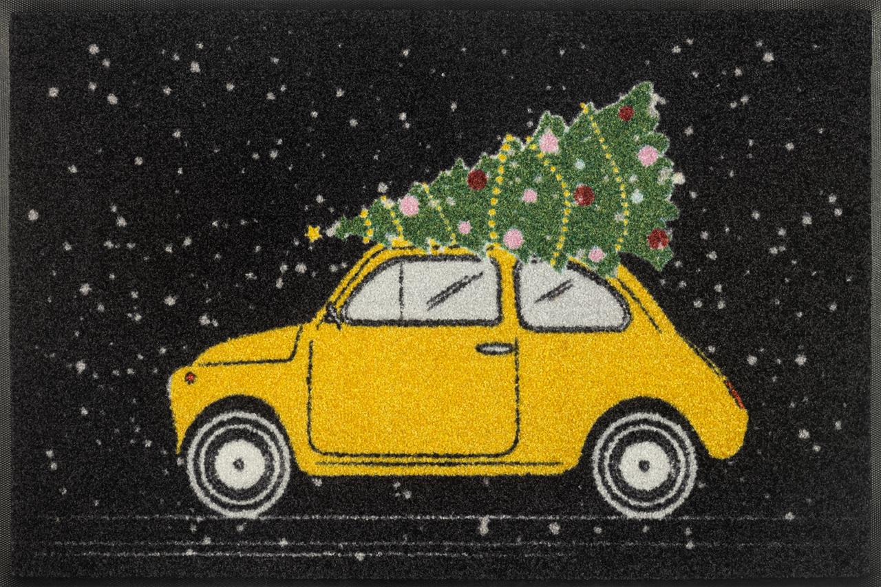 Auf der Fußmatte ist ein gelbes Auto zu sehen, das einen geschmückten Weihnachtsbaum auf dem Dach transportiert. Im Hintergrund fällt Schnee auf den schwarzen Untergrund.