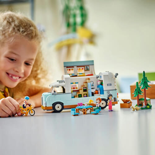 Ein Kind spielt mit einem LEGO-Wohnmobil-Set auf einem Tisch. Verschiedene kleine Figuren, ein Picknickplatz und ein Baum sind ebenfalls zu sehen.