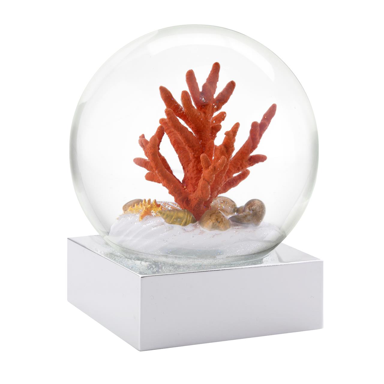 In der Schneekugel „Coral Sea“ befindet sich eine rote Koralle auf weißem Sand mit kleinen Muscheln. Die Glaskugel steht auf einem glänzenden, eckigen Sockel.