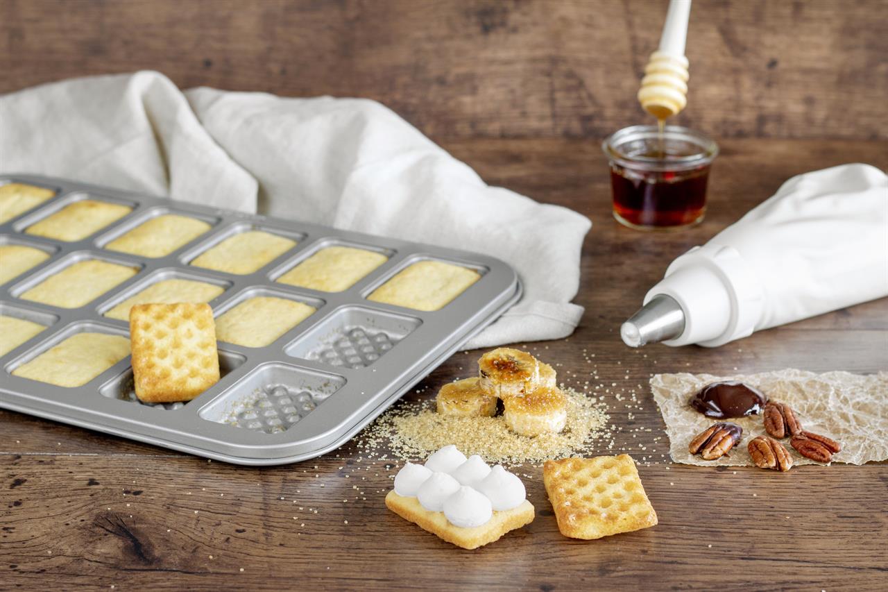 Das Bild zeigt ein Mini-Waffelblech mit frisch gebackenen kleinen Waffeln. Im Vordergrund liegen verschiedene Zutaten wie Nüsse, Schokolade und eine Dekorier-Spritze.