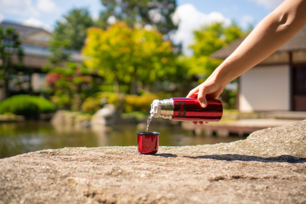 Eine Person gießt Wasser aus einer roten Thermosflasche in einen kleinen Becher. Im Hintergrund befindet sich ein Garten mit Bäumen und einem Teich.