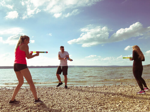 Drei Personen spielen am Ufer eines Sees mit Wasserpistolen. Es ist sonnig und der Himmel ist mit Wolken bedeckt.