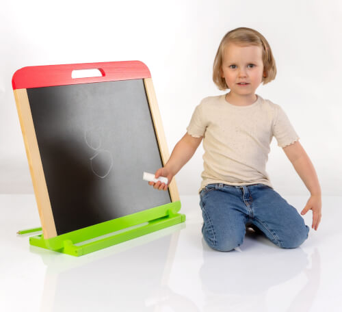 Ein kleines Kind sitzt auf dem Boden neben einer Tafel und hält ein Stück Kreide in der Hand. Auf der Tafel sind einfache Kreidezeichnungen zu sehen.