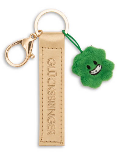 Auf dem Bild ist ein Schlüsselanhänger mit einem goldenen Lederband, auf dem "GLÜCKSBRINGER" steht, zu sehen. Daran hängt ein grünes, plüschiges Kleeblatt mit einem lächelnden Gesicht.
