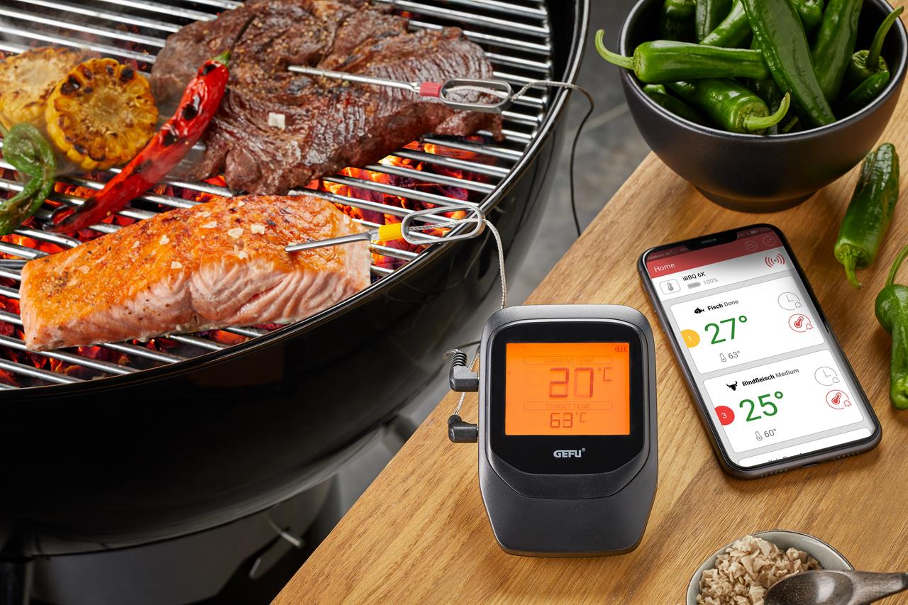 Auf dem Bild ist ein Grill mit Fleisch, Fisch und Gemüse zu sehen, in die Temperatursonden gesteckt sind. Daneben stehen das Grill- und Bratenthermometer Control+ 6 Kanal sowie ein Smartphone mit einer App, die die aktuellen Temperaturen anzeigt.