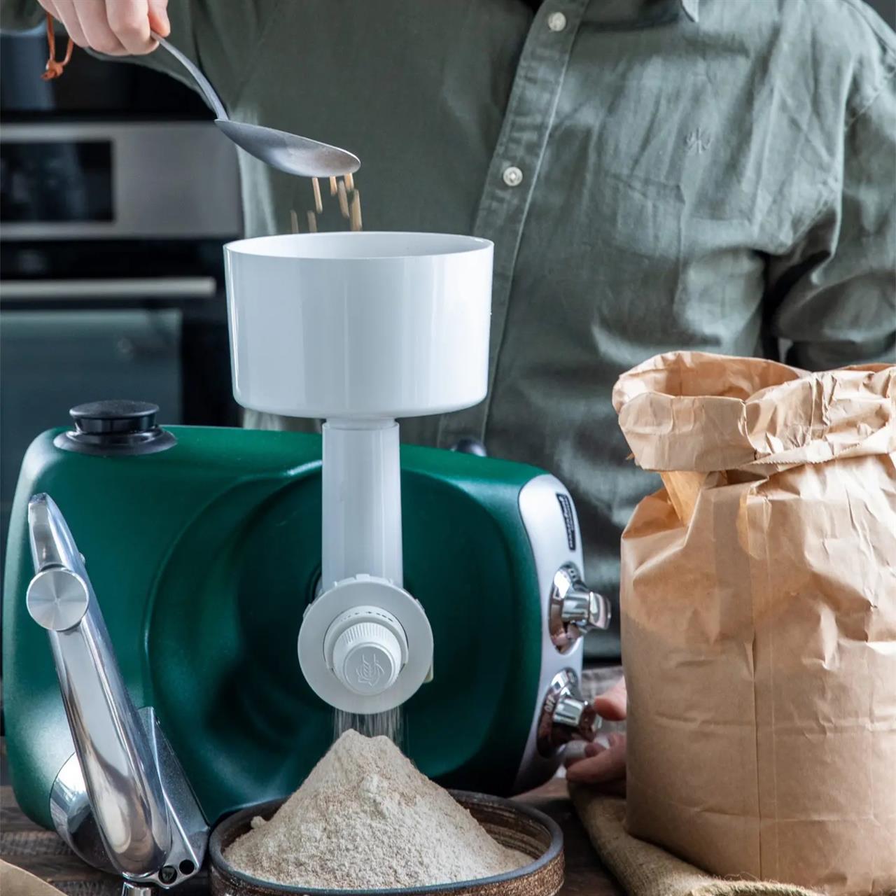 Eine Person gibt mit einem Löffel Getreide in die Getreide- und Kaffeemühle. Frisch gemahlenes Mehl fällt in eine Schale darunter.