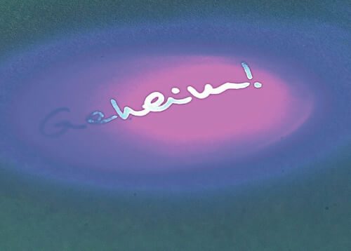 Auf dem Bild ist das Wort "Geheim!" zu sehen, das auf einem Untergrund geschrieben wurde. Es leuchtet im Schein eines UV-Lichts in lila und blau.