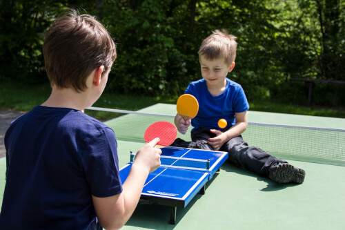 Zwei Jungen spielen Tischtennis an einem kleinen Tisch im Freien. Beide tragen blaue T-Shirts und wirken konzentriert auf das Spiel.