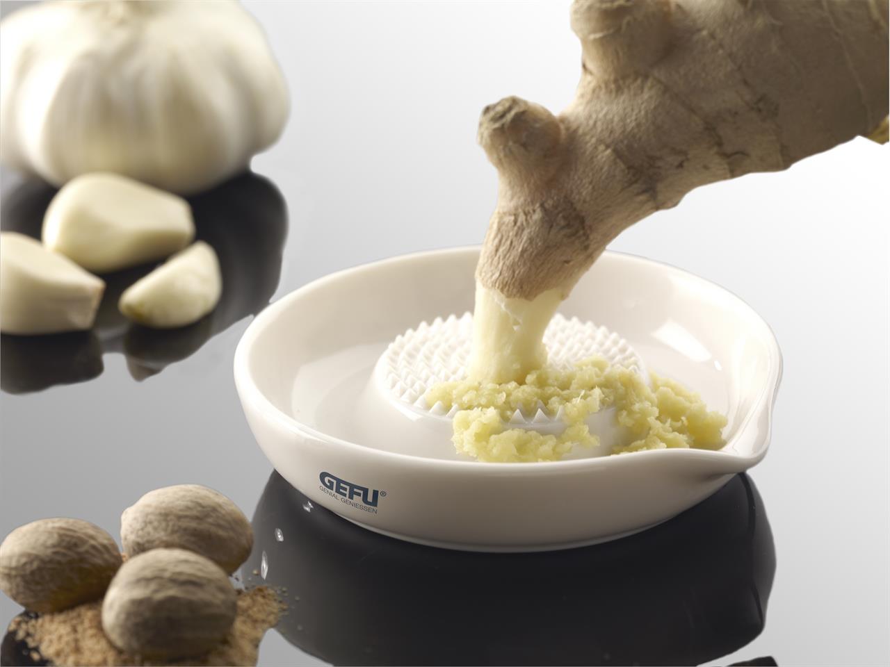 Das Bild zeigt eine Ingwerreibe aus Keramik, auf der frischer Ingwer gerieben wird. Im Hintergrund liegen Knoblauchzehen und Muskatnüsse auf einer glänzenden Oberfläche.