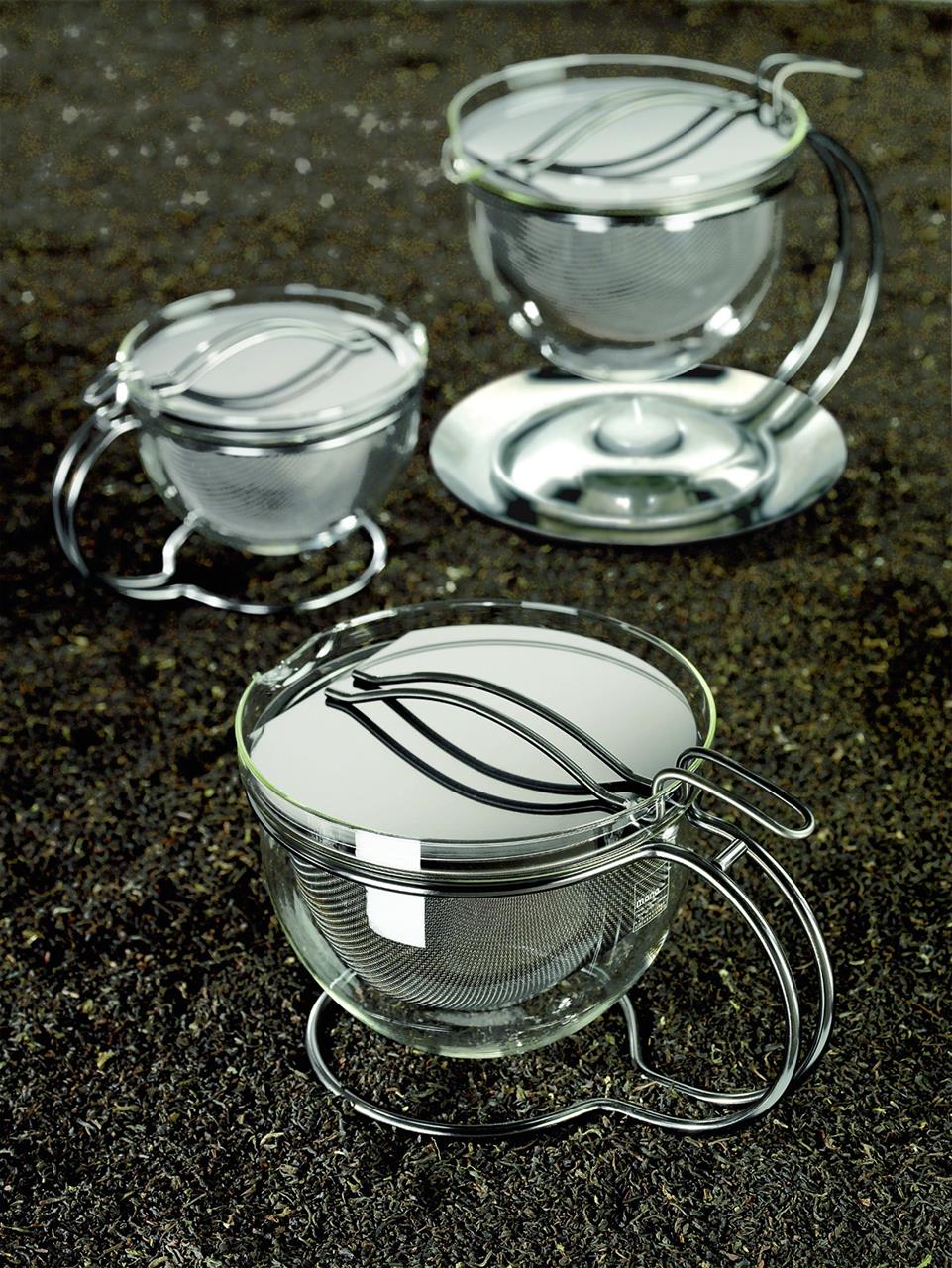 Das Bild zeigt drei runde Teekannen mit Griff aus Glas und Metall. Sie stehen auf einer dunklen Oberfläche, vermutlich losem Tee.