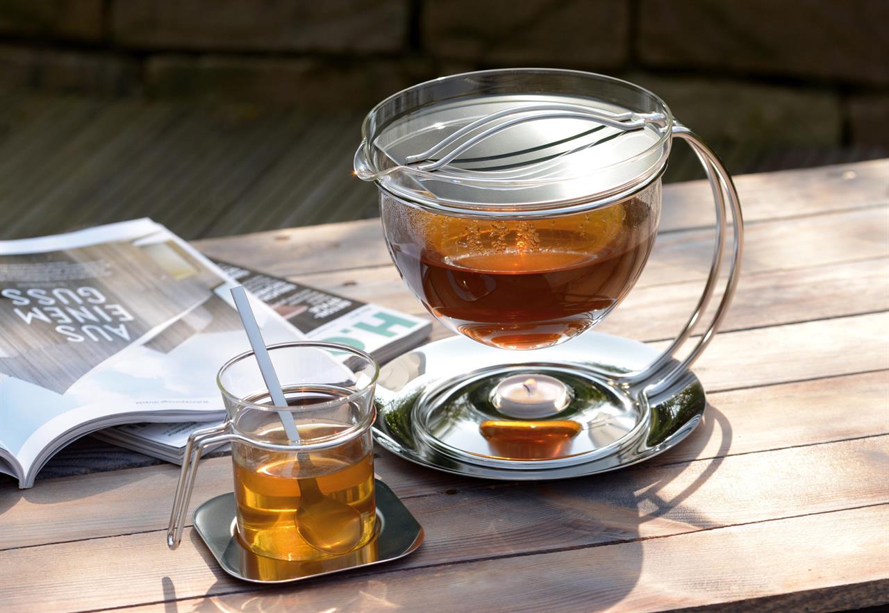 Auf dem Bild ist die Teekanne Filio 1,5L mit frisch aufgebrühtem Tee zu sehen. Daneben steht ein Glas mit Tee und ein aufgeschlagenes Magazin auf einem Holztisch.