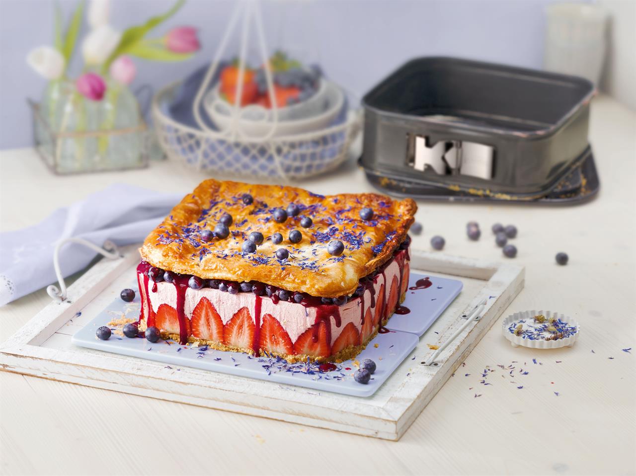 Im Vordergrund steht ein quadratischer Kuchen mit Erdbeeren, Sahne, Heidelbeeren und Blätterteig. Im Hintergrund ist die quadratische Springform "La Forme Plus-Neu 24x24cm" zu sehen.