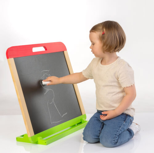 Ein kleines Mädchen sitzt auf dem Boden und wischt eine Tafel sauber. Sie hat ein Stück Kreide oder einen Schwamm in der Hand und trägt ein beigefarbenes T-Shirt und eine blaue Jeans.