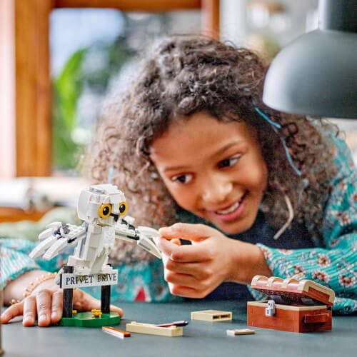 Ein Kind spielt lächelnd mit einem LEGO-Set auf einem Tisch. Im Vordergrund ist eine weiße Eule mit einem Schild "Privet Drive" und eine kleine Schatztruhe zu sehen.