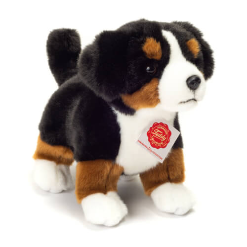 Das Bild zeigt einen Plüschhund mit schwarzem, braunem und weißem Fell. Am Hals trägt er ein rotes Etikett mit einem Logo.