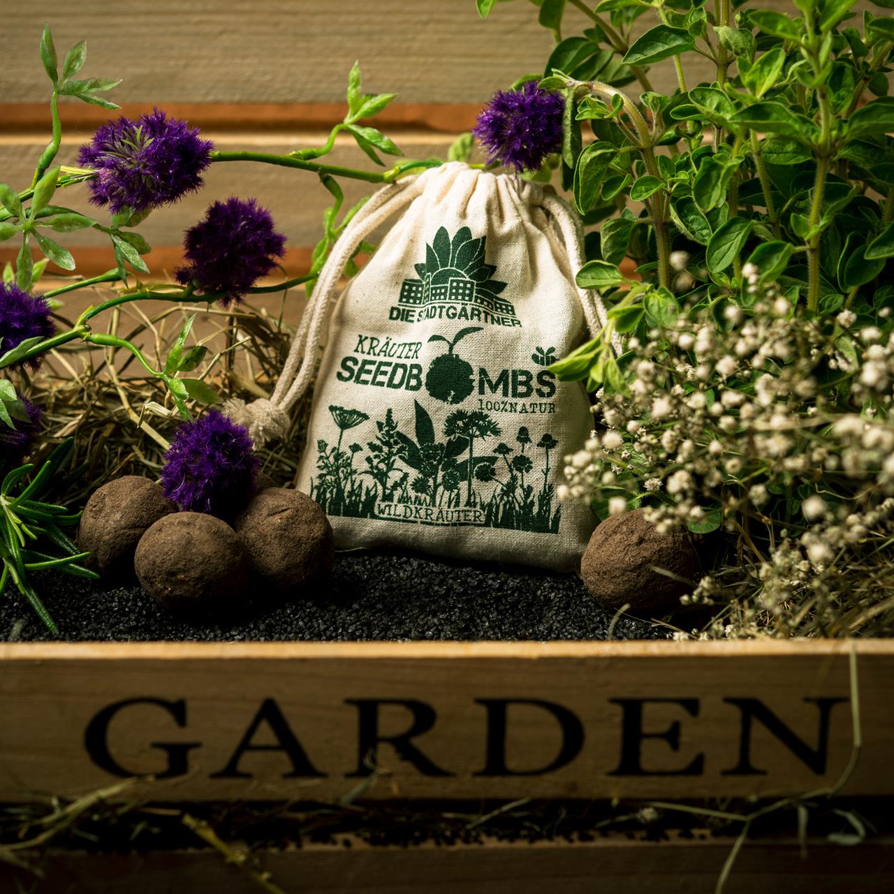 Auf dem Bild ist ein Jutebeutel mit der Aufschrift "Wildkräuter Samenbombe" zu sehen, umgeben von Pflanzen und Samenbomben. Der Beutel steht in einer Holzkiste mit der Aufschrift "GARDEN".