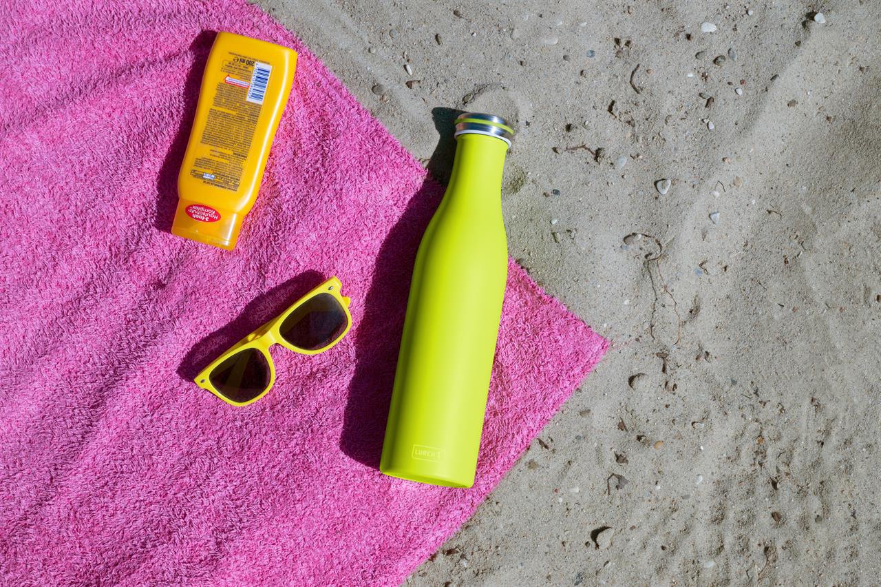 Auf dem Bild liegt eine Isolier-Flasche aus Edelstahl in der Farbe Fresh Green auf einem pinken Handtuch im Sand. Daneben befinden sich eine gelbe Sonnenbrille und eine gelbe Sonnencreme.
